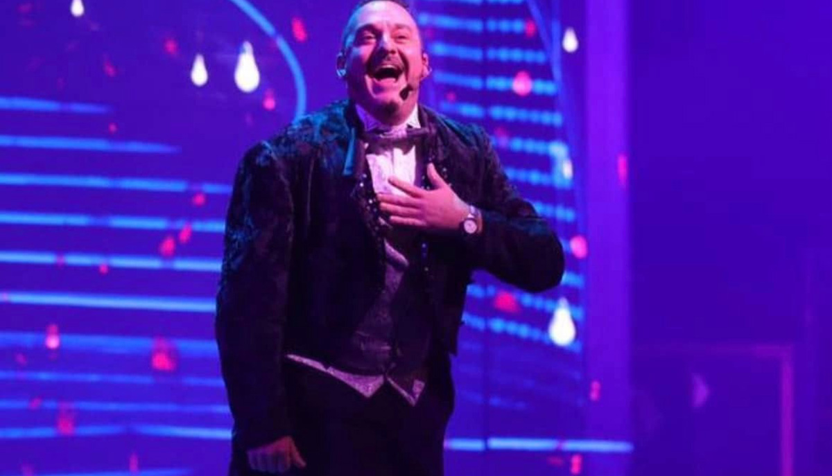A man in a tuxedo is performing on stage with vibrant purple lighting and a joyful expression.