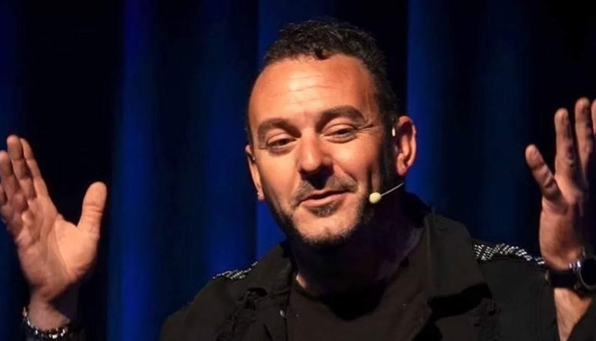 A man is speaking on stage with a microphone, gesturing with both hands against a dark blue background.