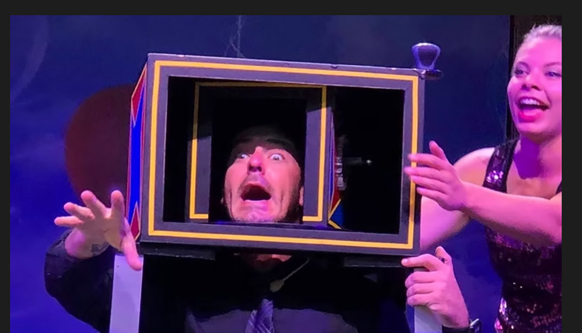 A person is playfully making a surprised face while their head is inside a colorful magician's box, and another person is smiling next to them.
