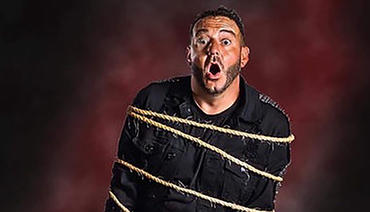 A man in a black outfit is tied with ropes and has a surprised expression on his face against a dark, blurry background.