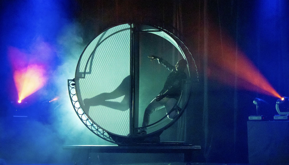 A performer is dramatically silhouetted inside a large circular structure on stage, surrounded by colorful lights and smoke effects.