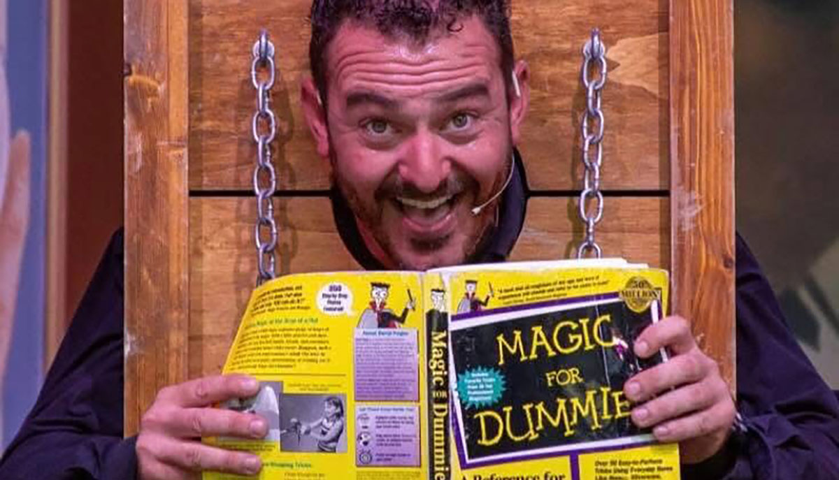 A man with a headset is humorously looking through a wooden restraint while holding and reading 