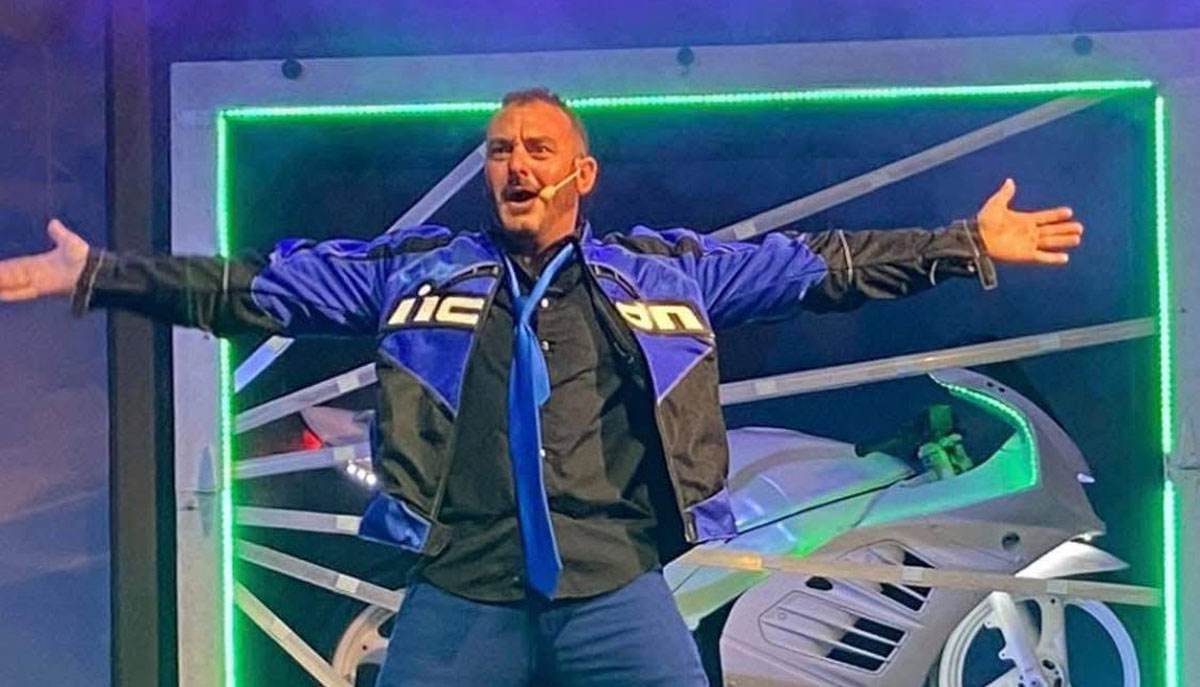 A man wearing a blue and black jacket with a headset microphone stretches his arms wide open on stage, with a futuristic motorcycle-like object in the background.