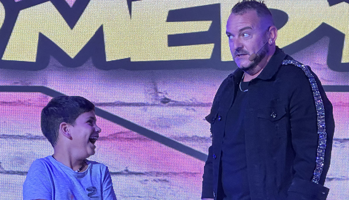 A boy and a man are on stage, with the boy laughing and the man looking amused or surprised, wearing a microphone headset.