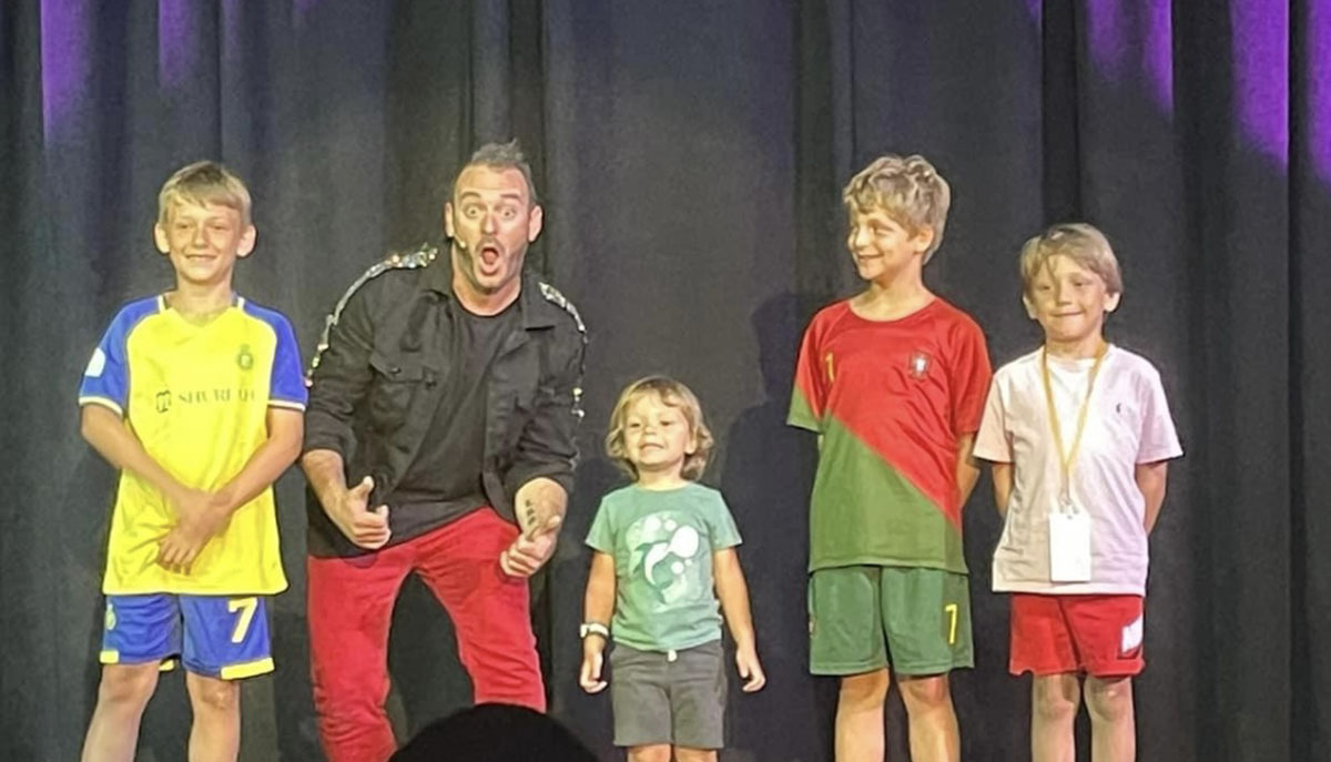 A man and four children are standing in a row on stage, with the man making a playful expression.
