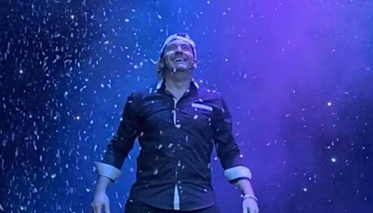 A person is joyfully standing under a snowfall-like effect, surrounded by vibrant blue and purple lighting.