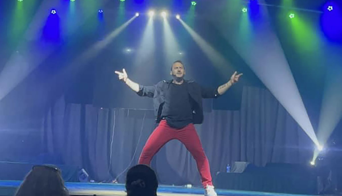 A performer in red pants and a black jacket strikes a dynamic pose on stage under colorful spotlights.