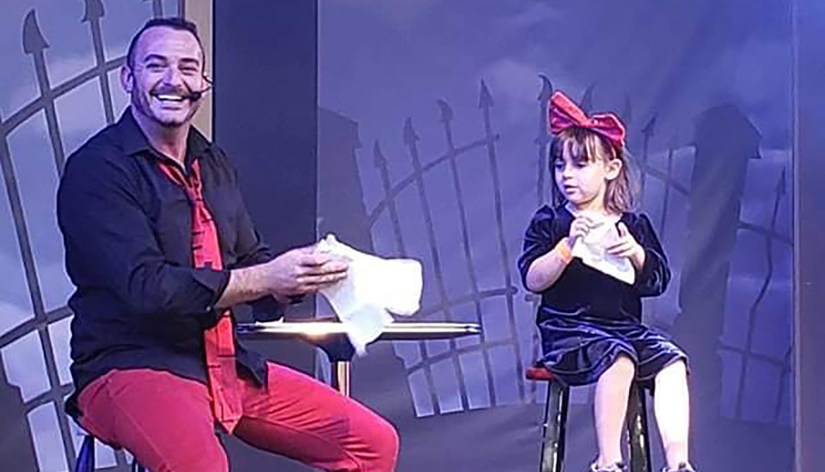 A man and a young girl, both smiling, are on stage performing a magic trick involving paper.