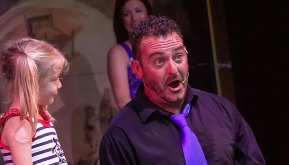 A man with a microphone and a surprised expression is interacting with a smiling young girl on stage.