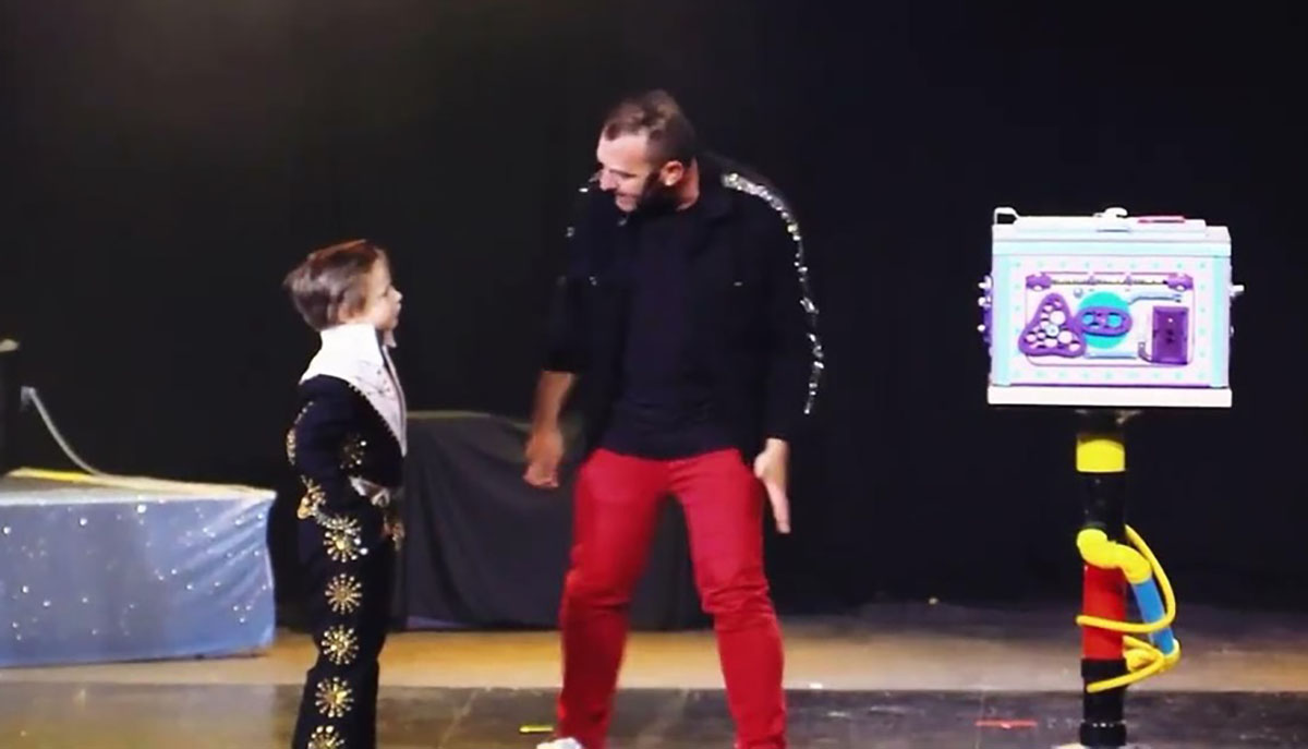 A child and an adult are performing a magic act on stage with a colorful box.