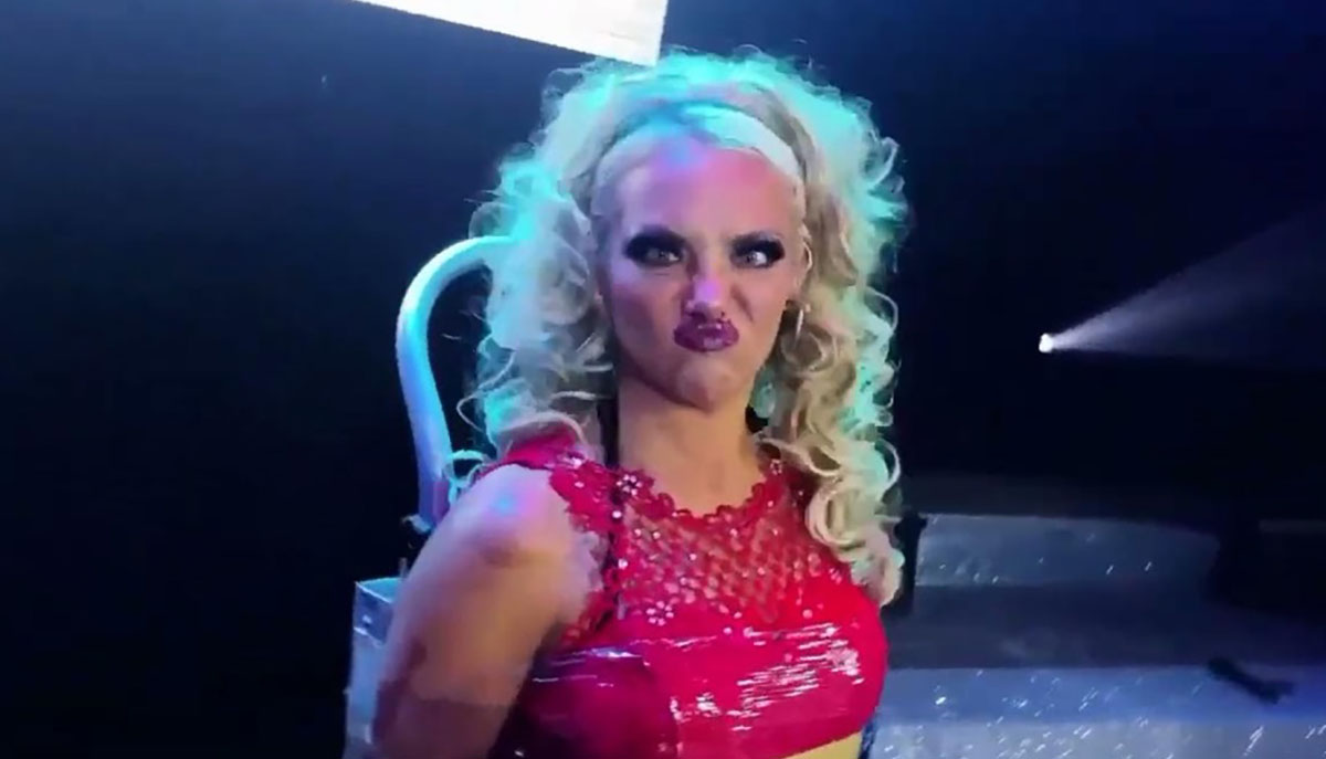 A person with curly blonde hair, wearing a shiny red outfit, is making a playful, pouty face on stage.