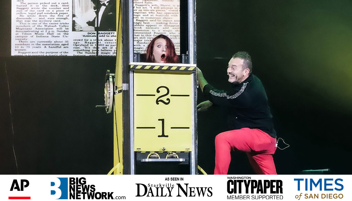 A woman looks surprised inside a box marked with numbers, while a smiling man interacts with the setup during a likely magic performance.